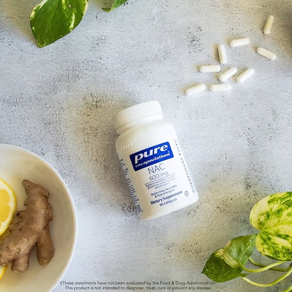 A clean, top-down shot of a Pure Encapsulations NAC 600mg bottle surrounded by fresh lemon, ginger, and supplement capsules on a textured concrete surface.